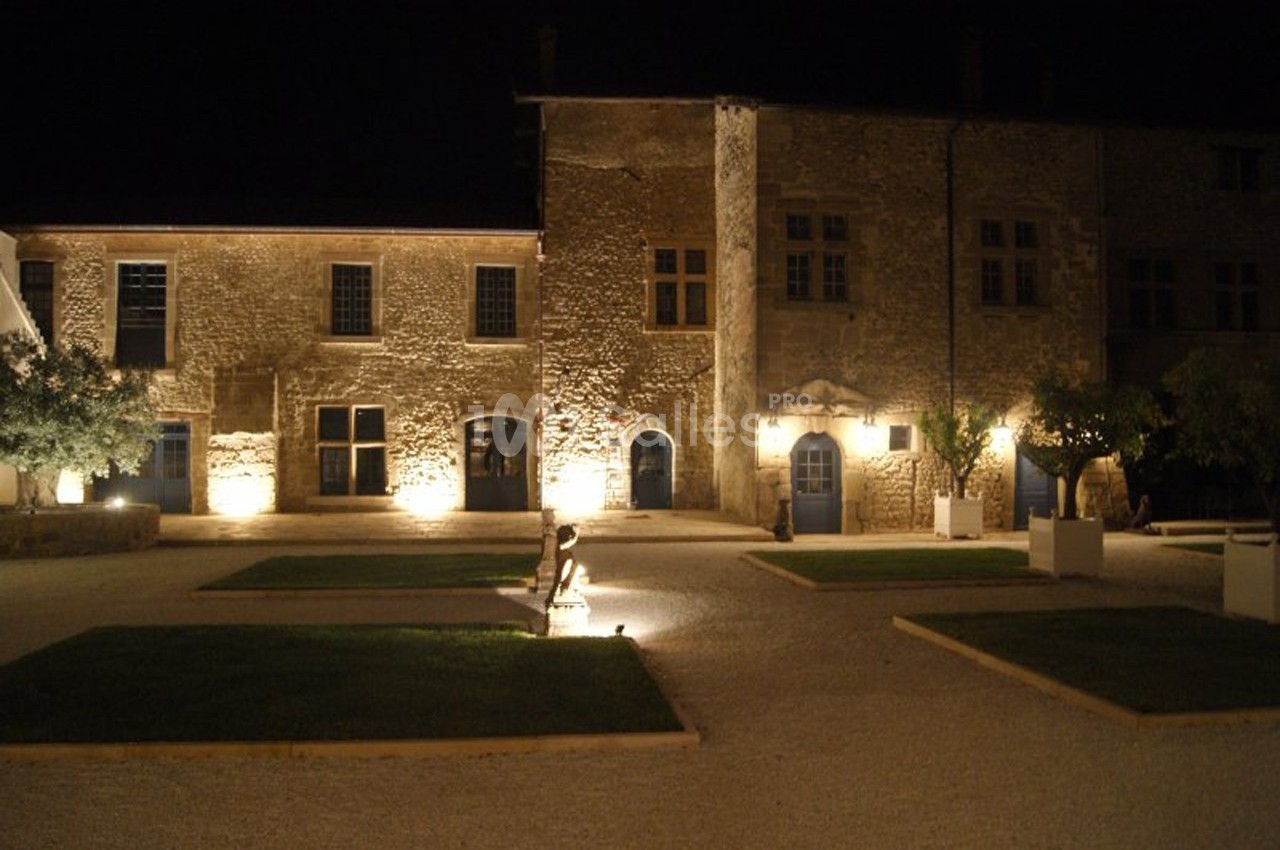 Façade en pierre d'un bâtiment ancien éclairé la nuit, entourée de pelouses et d'allées gravillonnées.