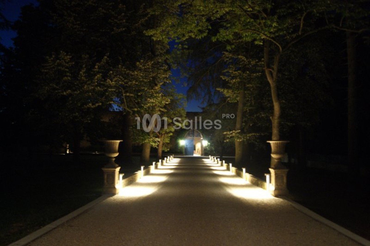Allée illuminée bordée d'arbres et de lampadaires, menant à une porte éclairée dans un cadre nocturne.
