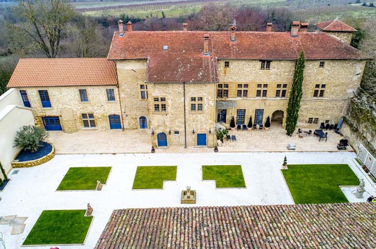 Vue aérienne d'un bâtiment en pierre avec toit en tuiles, cour aménagée en pelouses et allées gravillonnées.