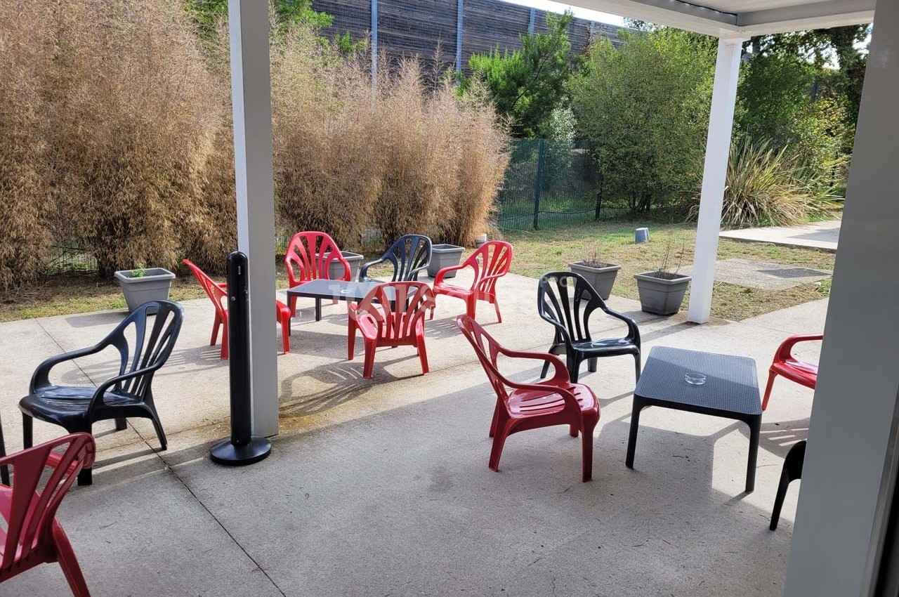Terrasse avec tables et chaises en plastique rouge et noir, entourée de végétation et protégée par un toit.