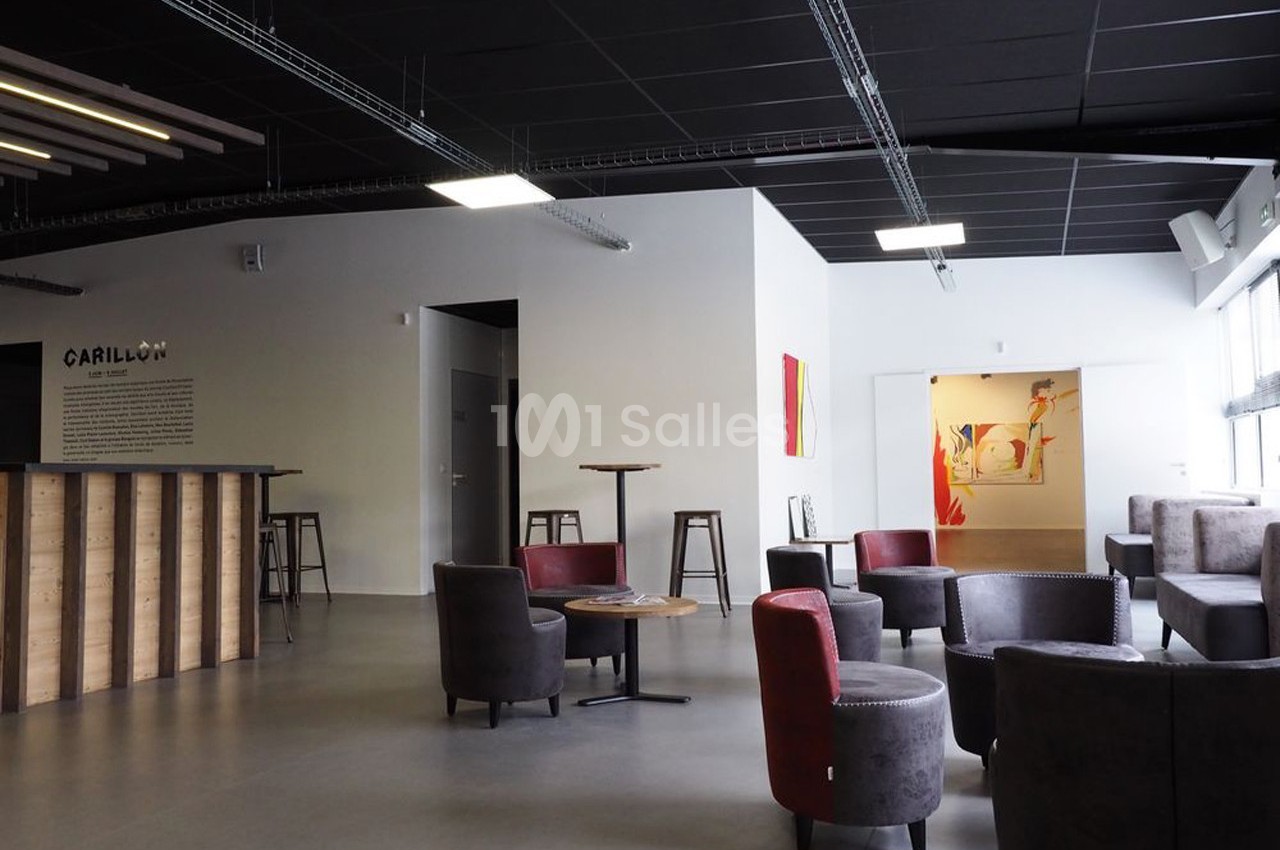 Espace lounge moderne avec fauteuils, tables basses, comptoir en bois et murs décorés, éclairé par des fenêtres.