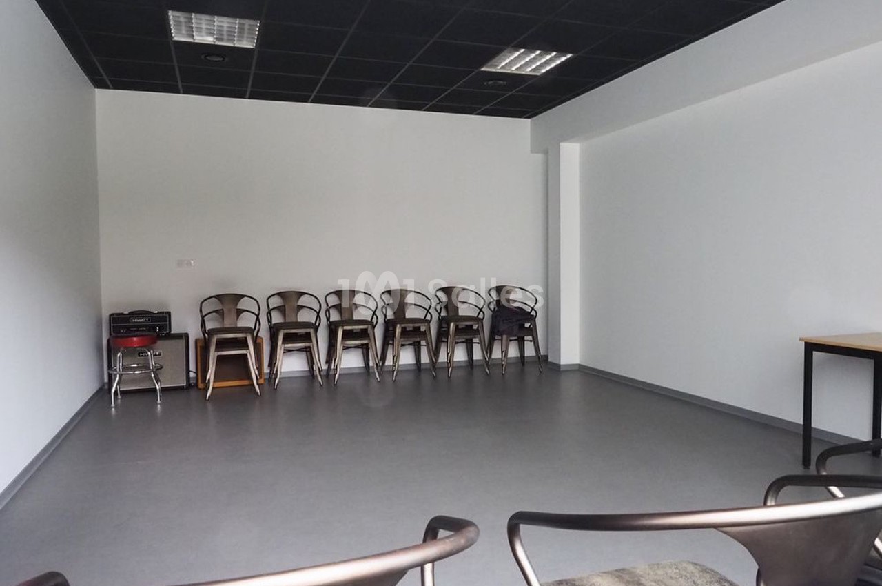 Salle vide avec des chaises alignées contre un mur blanc, sol gris et plafond noir avec éclairage fluorescent.