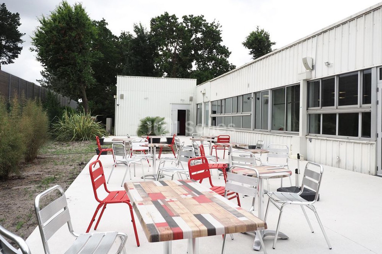 Terrasse extérieure avec tables et chaises en métal, entourée de végétation et attenante à un bâtiment blanc.