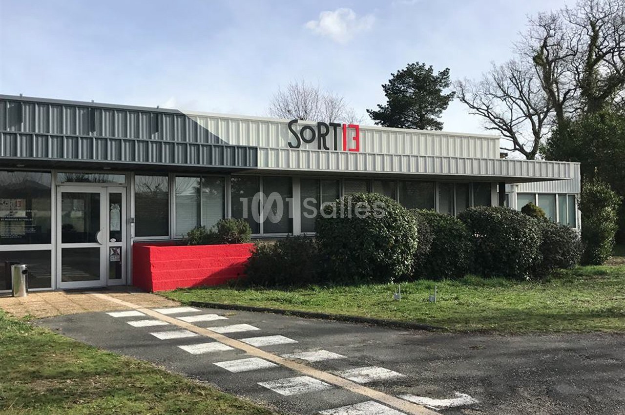 Façade d'un bâtiment moderne avec le logo ’SORTIE’, entouré de verdure et d'un passage piéton au premier plan.