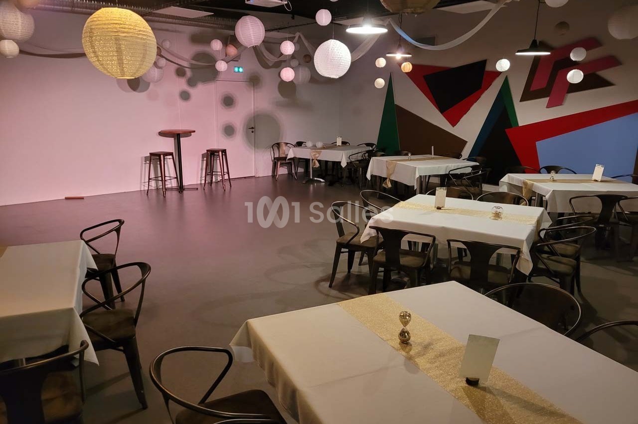 Salle décorée avec des tables dressées, des chaises noires, des lampions suspendus et un mur coloré en arrière-plan.