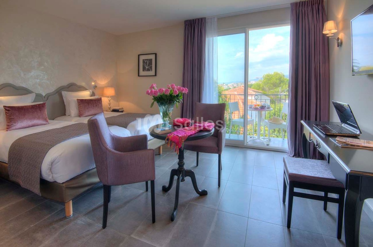 Chambre lumineuse avec lit double, table et chaises, balcon donnant sur un paysage arboré, bureau avec ordinateur portable.