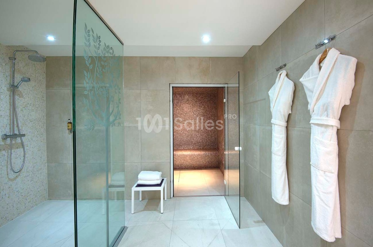 Salle de bain moderne avec douche à l'italienne, peignoirs accrochés et accès à un hammam en arrière-plan.