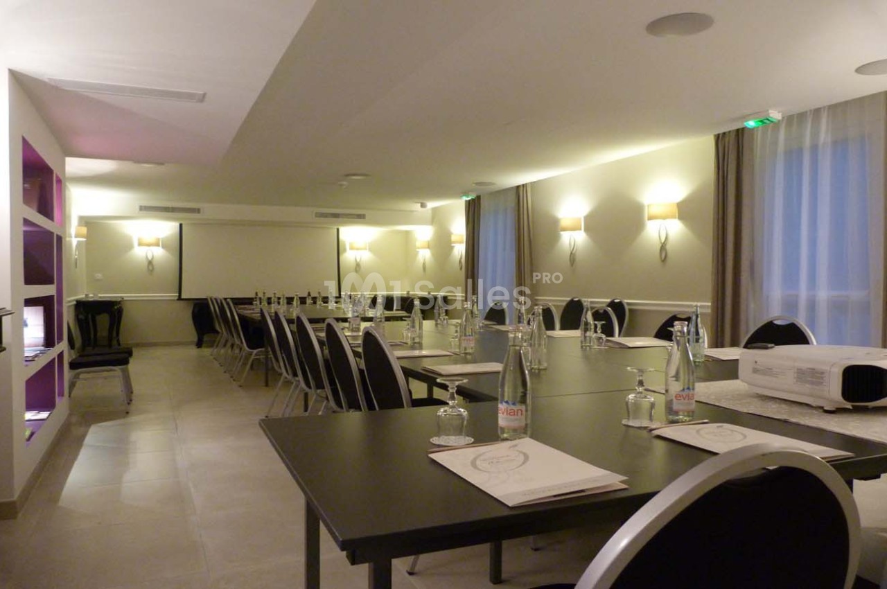Salle de réunion lumineuse avec tables noires disposées en U, bouteilles d'eau, documents et vidéoprojecteur.