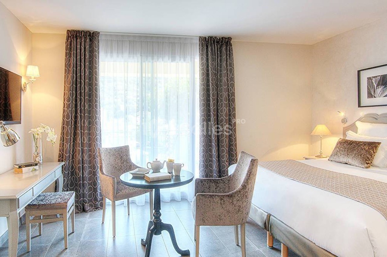 Chambre lumineuse avec lit double, table ronde, deux fauteuils, bureau, télévision murale et grande fenêtre avec rideaux.