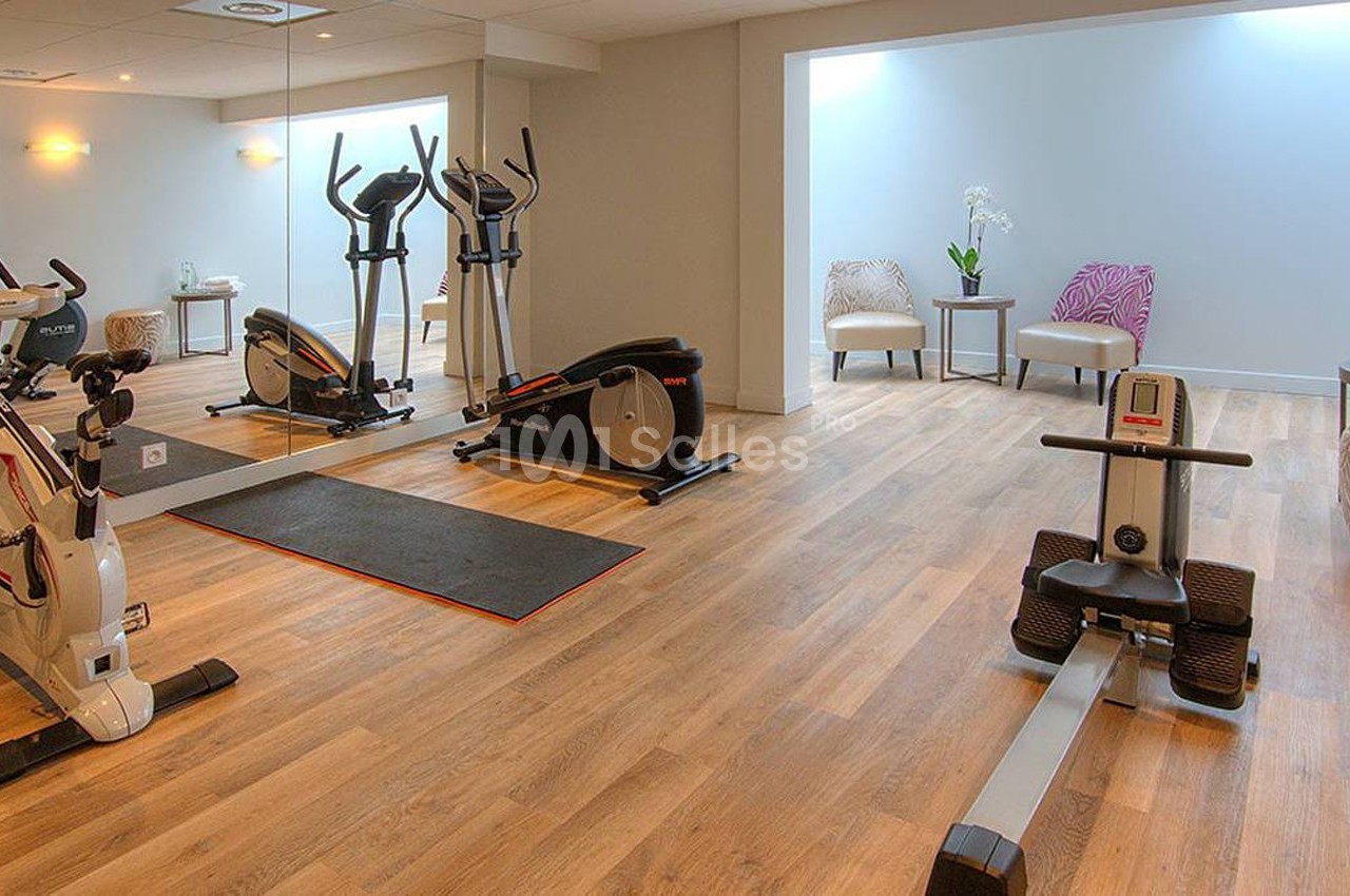 Salle de sport lumineuse avec vélos d'exercice, rameur, miroir mural et coin détente avec fauteuils.