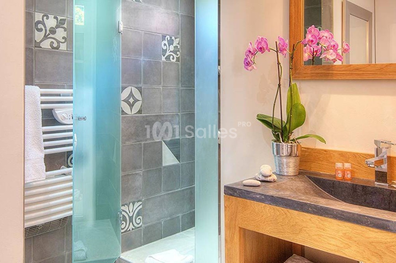 Salle de bain avec douche à porte vitrée, lavabo en bois avec miroir, orchidée et serviettes pliées.
