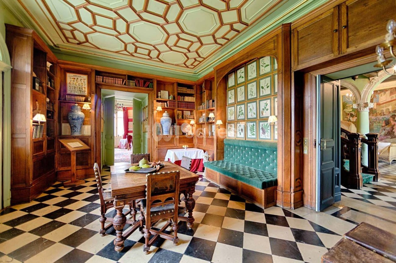 Bibliothèque avec boiseries, banquette verte, table en bois, sol en damier noir et blanc, et plafond à motifs géométriques.