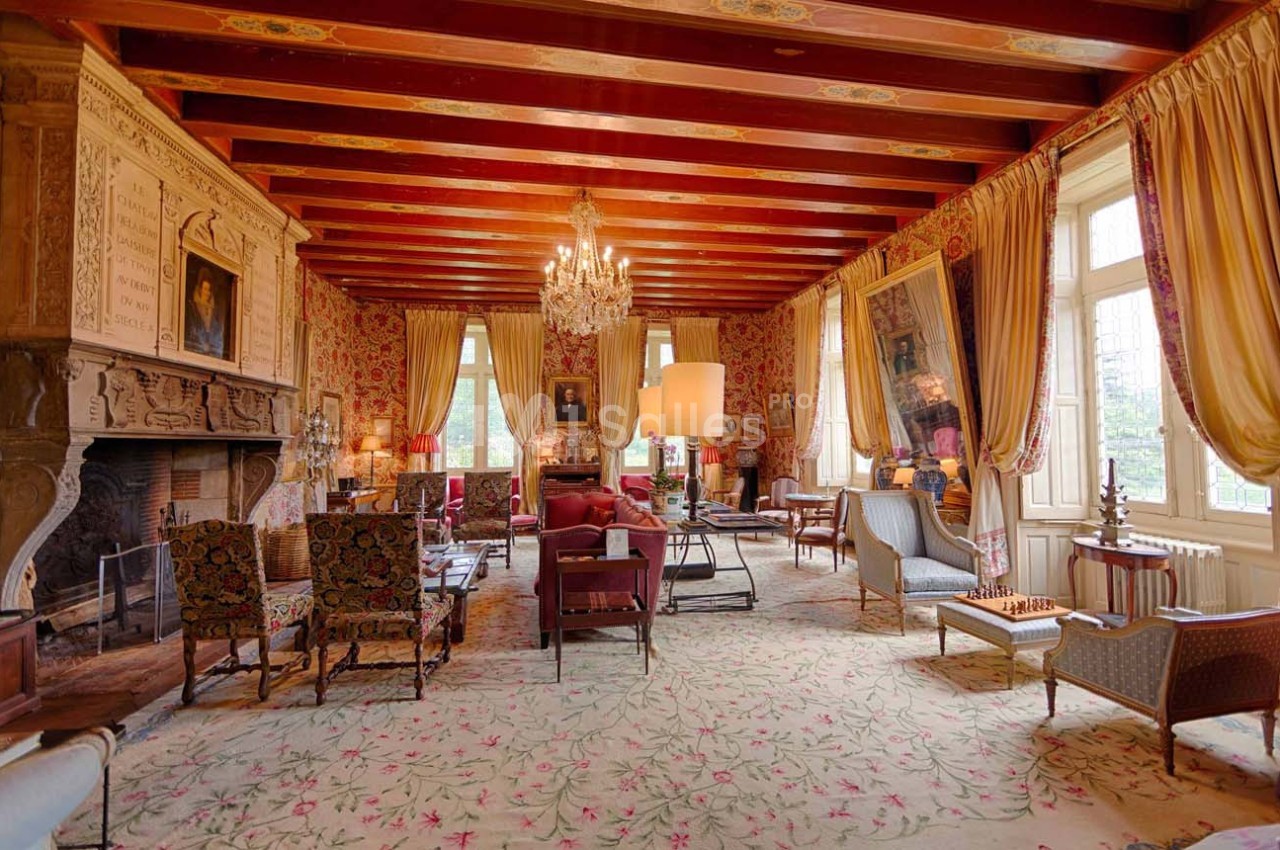 Salon élégant avec poutres apparentes, cheminée sculptée, lustre en cristal, tapis floral et mobilier classique.