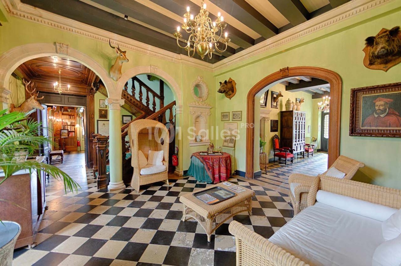Intérieur d'un salon avec sol en damier, mobilier en osier, trophées de chasse et un escalier en bois visible au fond.