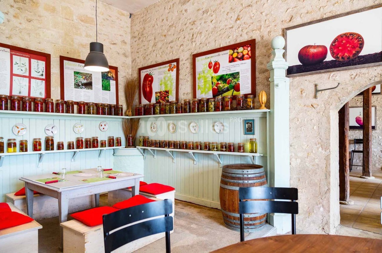Salle rustique avec des étagères remplies de bocaux de conserves, tables avec coussins rouges et panneaux explicatifs au mur.