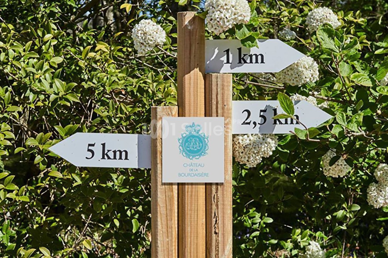Panneau en bois avec flèches indiquant des distances vers le Château de Bourbansais, entouré de végétation et de fleurs…