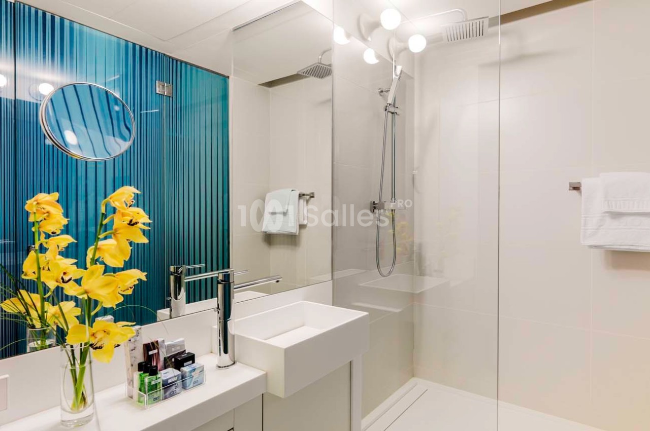 Salle de bain moderne avec douche vitrée, lavabo blanc, miroir rond, fleurs jaunes et mur bleu texturé.