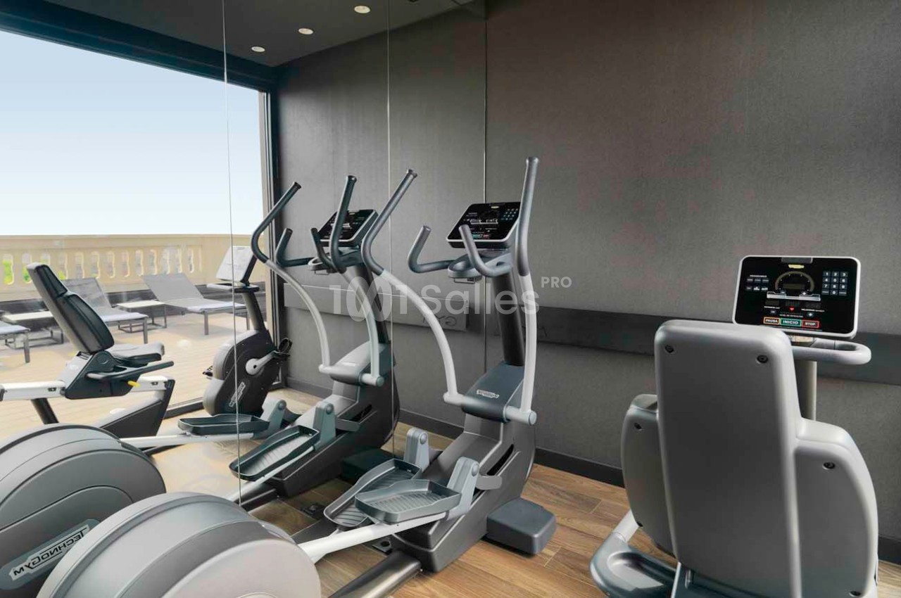 Salle de sport avec vélos elliptiques et tapis de course face à une baie vitrée donnant sur une terrasse extérieure.