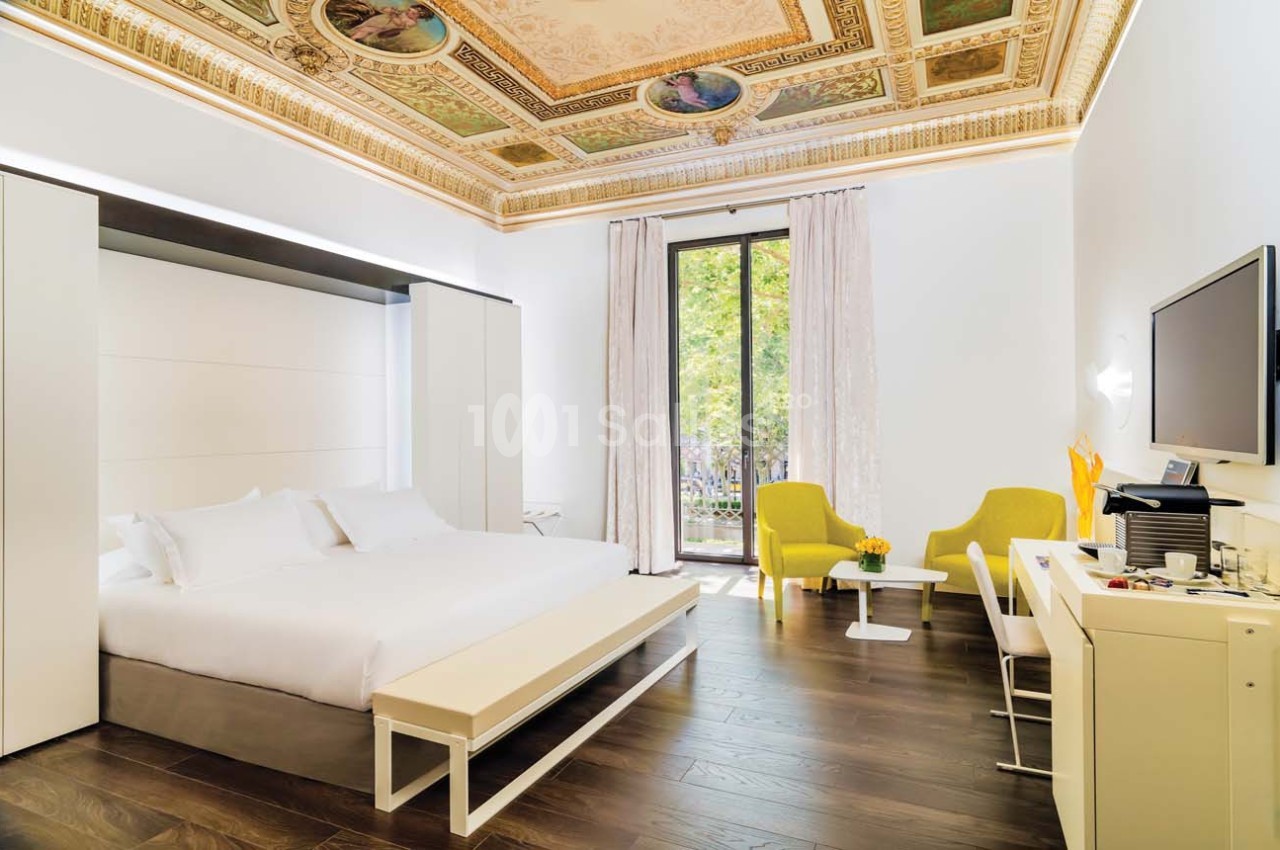 Chambre lumineuse avec lit double, mobilier moderne, plafond décoré, parquet foncé et vue sur un jardin par une grande…
