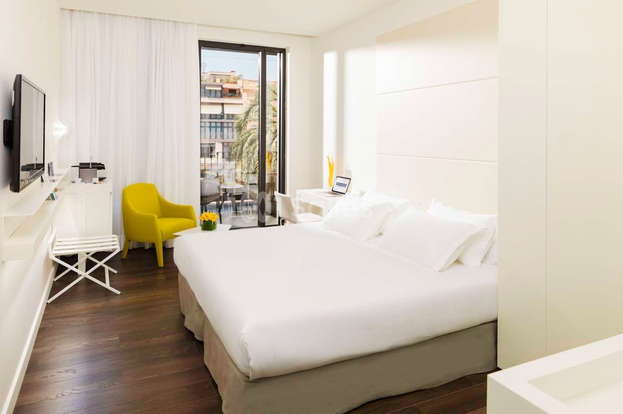 Chambre lumineuse avec lit double, fauteuil jaune, bureau, télévision murale et vue sur des bâtiments depuis une baie vitrée.