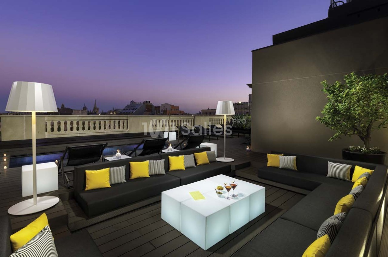Terrasse moderne avec mobilier sombre, coussins jaunes, table lumineuse et lampes, vue sur un ciel au crépuscule.