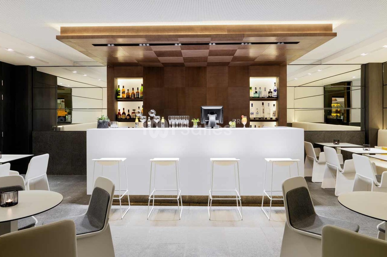 Bar moderne avec comptoir blanc, tabourets alignés, étagères éclairées contenant des bouteilles et verres en arrière-plan.