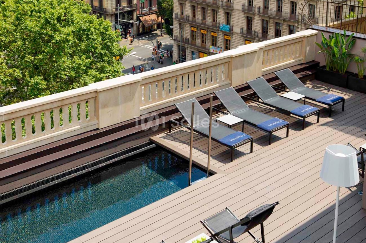 Terrasse avec piscine, transats alignés, plantes en pot et vue sur des bâtiments urbains et un arbre en arrière-plan.