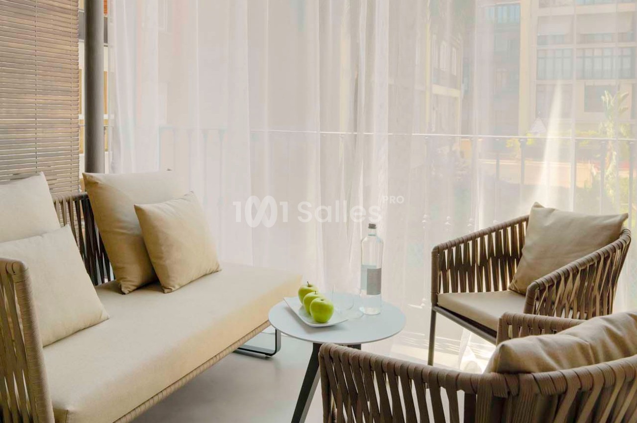 Salon extérieur avec canapé, fauteuils en rotin, table basse avec bouteille d'eau et pommes, devant un rideau translucide.