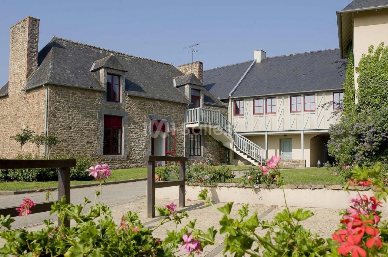 Maisons traditionnelles en pierre avec volets rouges et colombages, entourées de fleurs et d'une cour pavée.