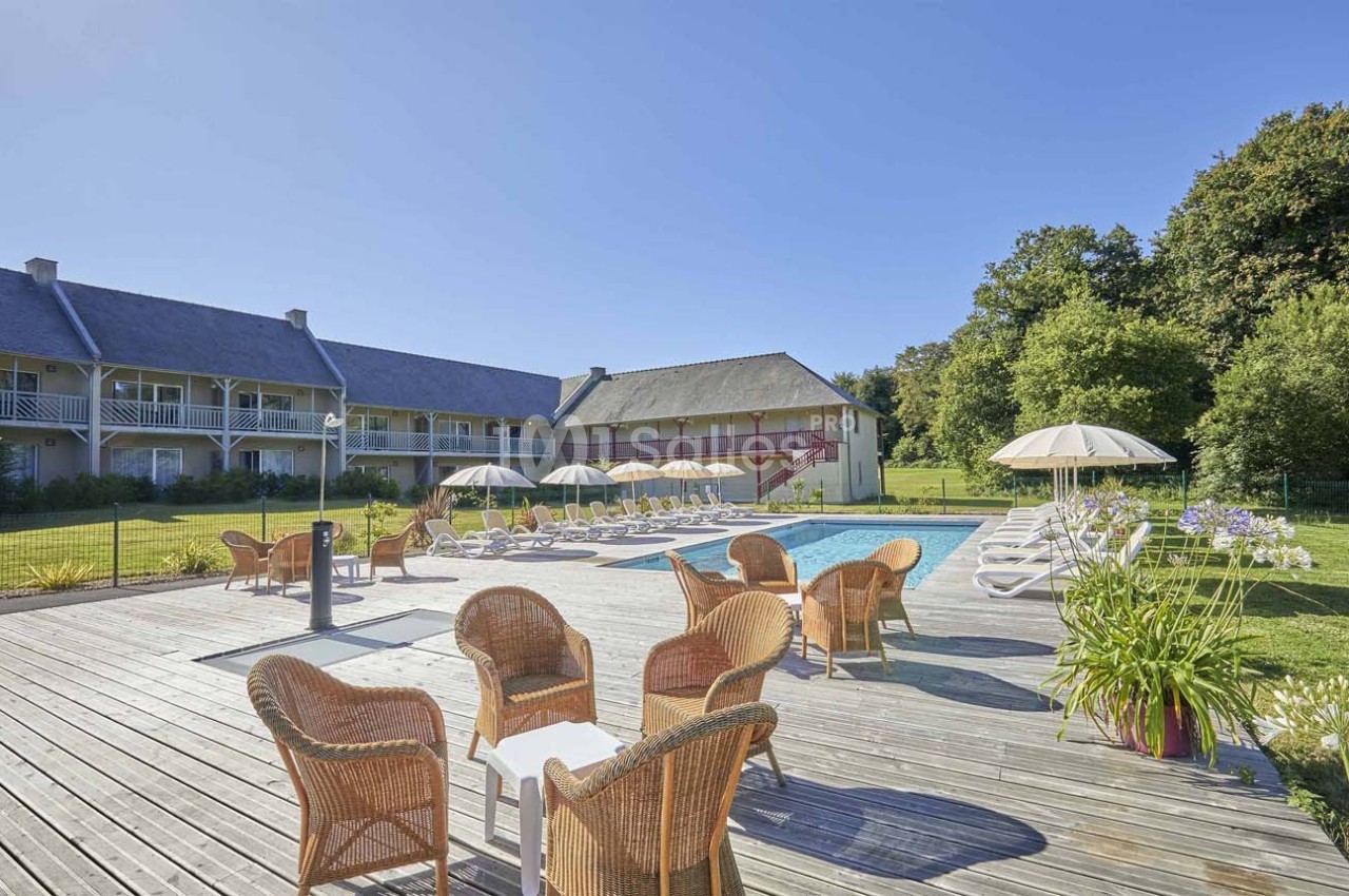 Terrasse en bois avec fauteuils en rotin, piscine extérieure entourée de transats et parasols, bâtiments en arrière-plan.