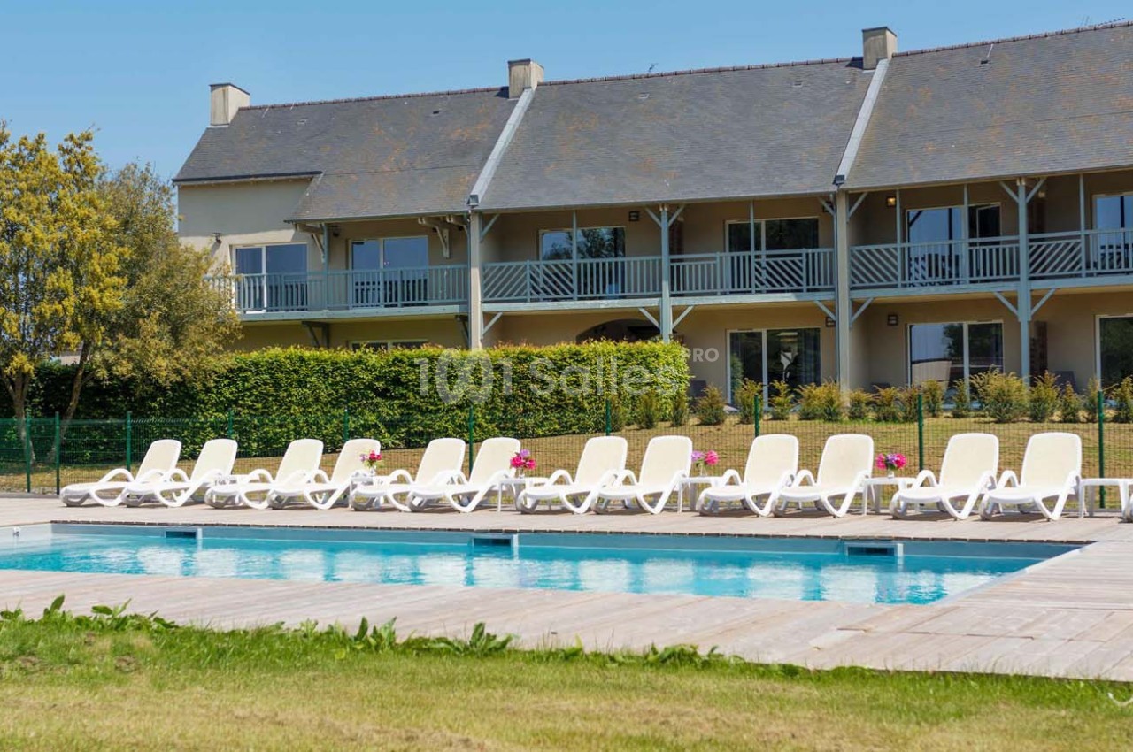 Piscine extérieure entourée de transats blancs devant un bâtiment résidentiel avec balcons et jardin.
