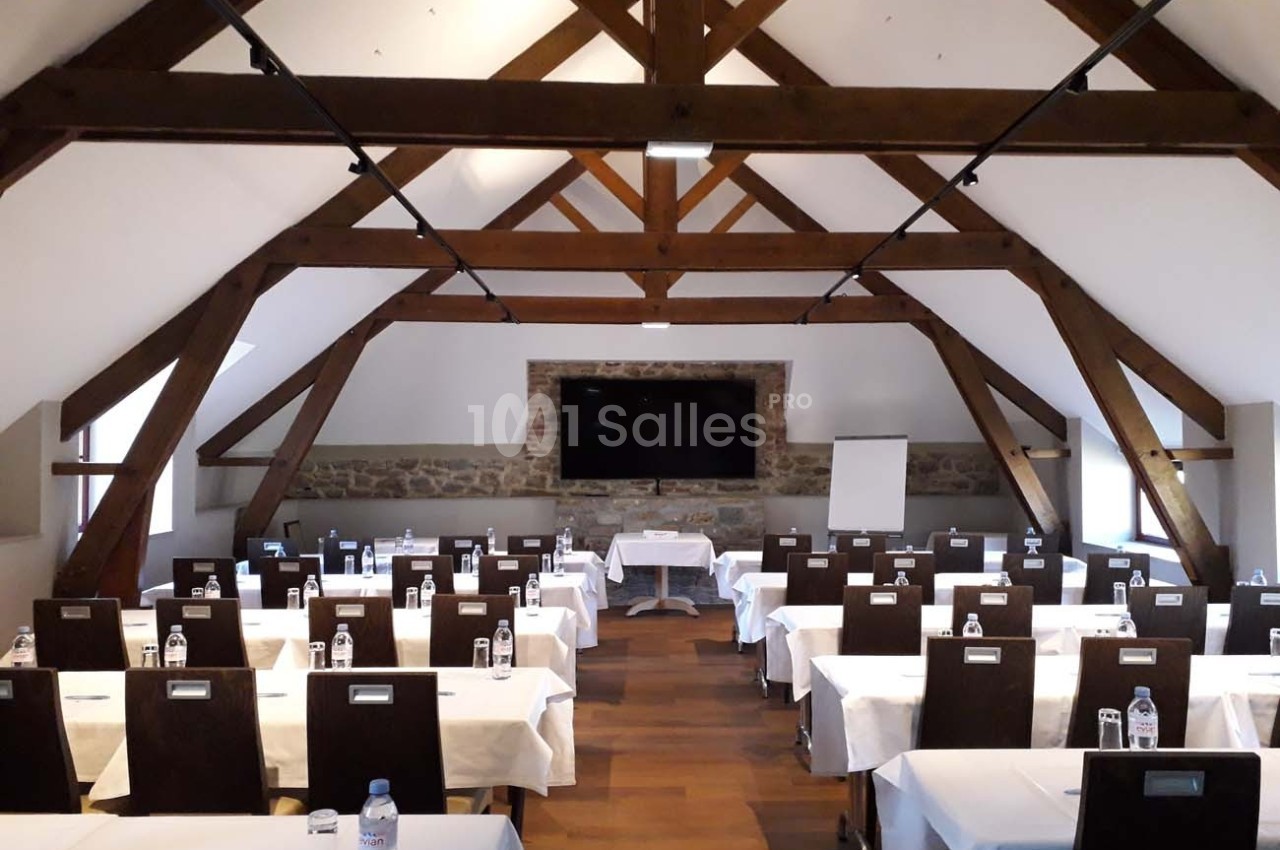 Salle de réunion avec poutres apparentes, tables alignées avec nappes blanches, écran et tableau blanc au fond.