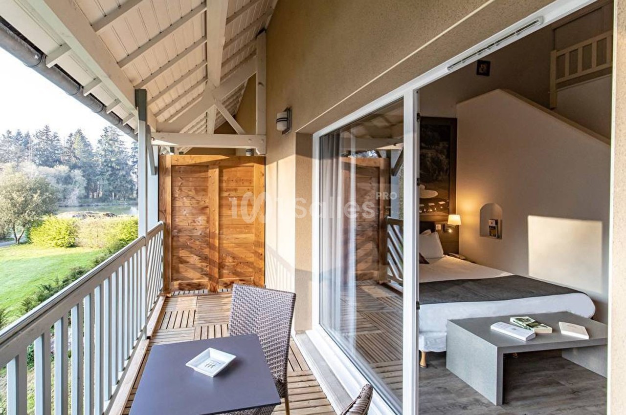 Balcon en bois avec table et chaises, vue sur un espace vert, donnant sur une chambre lumineuse avec un lit.
