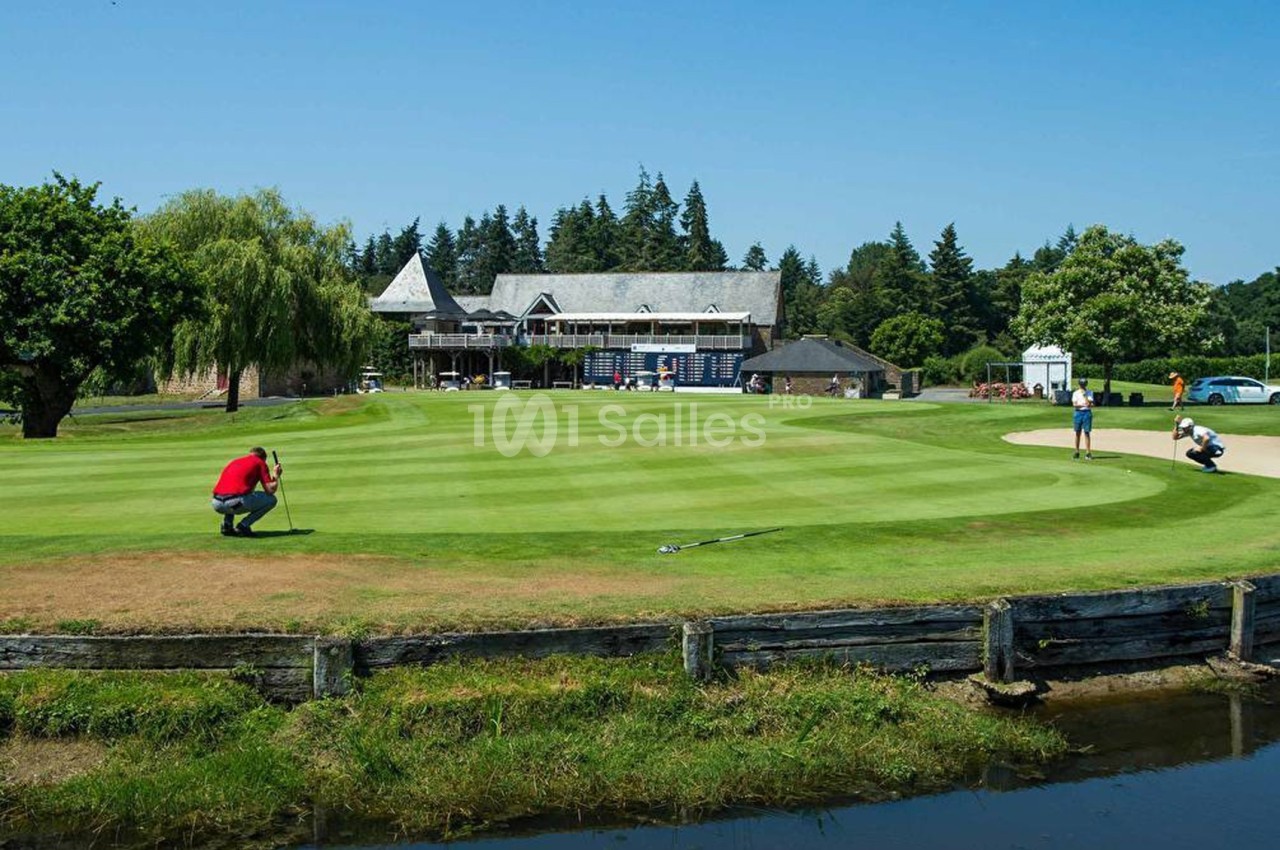 Un golfeur s'apprête à putter sur un green entouré de végétation, avec un club-house en arrière-plan.