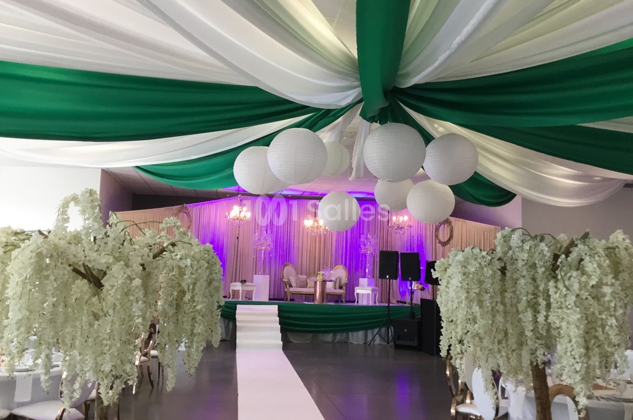 Salle de réception décorée avec des drapés verts et blancs, des lanternes suspendues et des arbres artificiels fleuris.