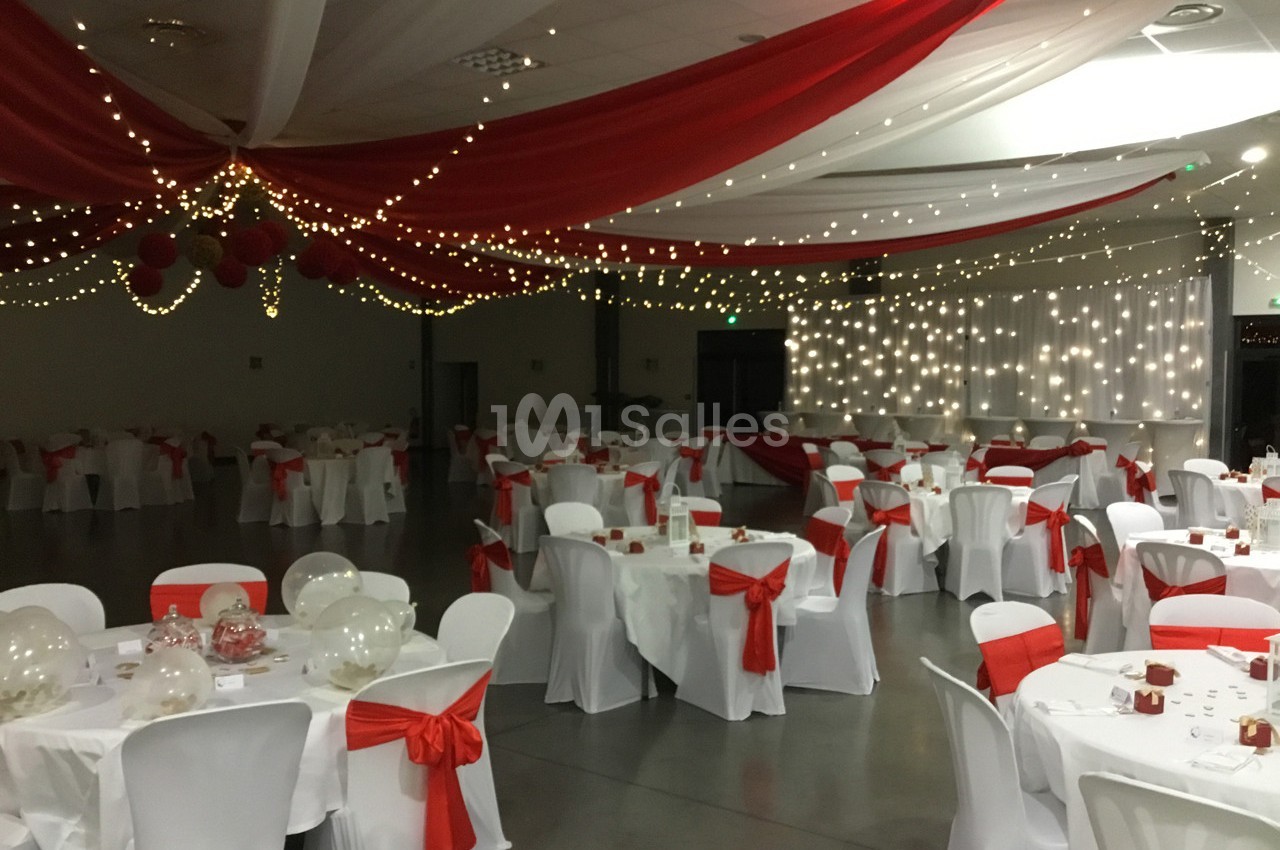 Salle de réception décorée avec des nappes blanches, des rubans rouges et des guirlandes lumineuses suspendues.