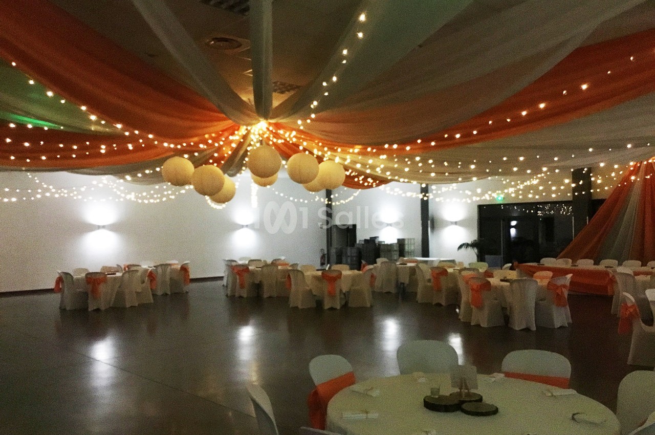 Salle de réception décorée avec des guirlandes lumineuses, des drapés orange et blanc, et des tables dressées.