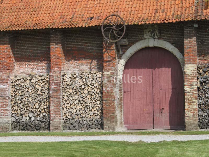 Façade d'une grange en briques avec porte en bois rouge, toit en tuiles et piles de bois empilées sur les côtés.
