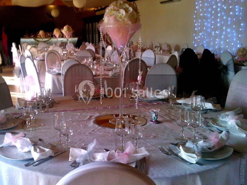 Salle de réception décorée avec des tables rondes, nappes blanches, serviettes roses et centre de table floral.
