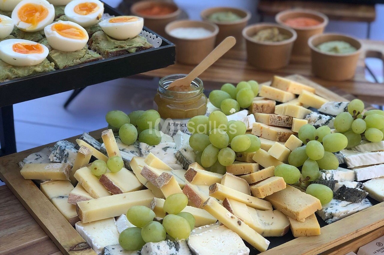 Plateau de fromages variés avec raisins verts, œufs durs et tartines d'avocat, accompagné de sauces en arrière-plan.