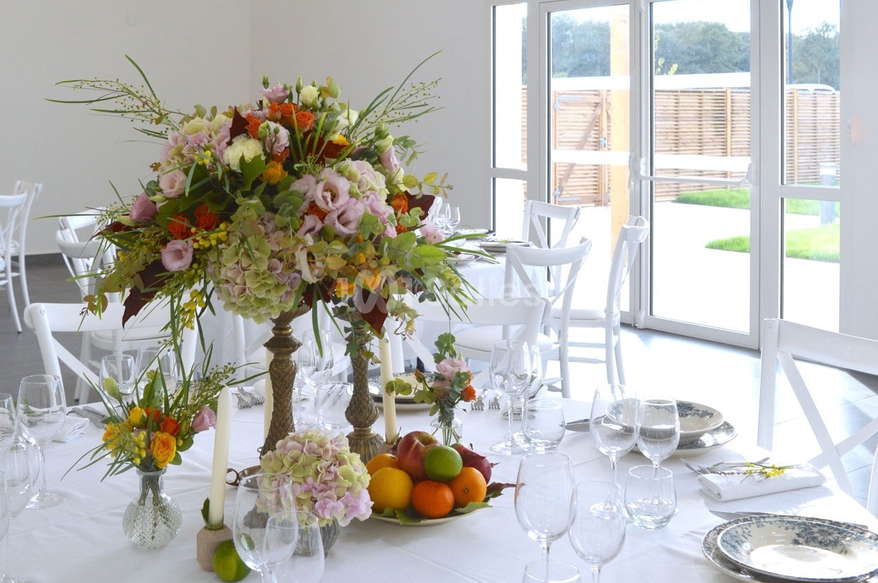 Centre de table floral avec fruits sur une table dressée dans une salle lumineuse aux grandes baies vitrées.