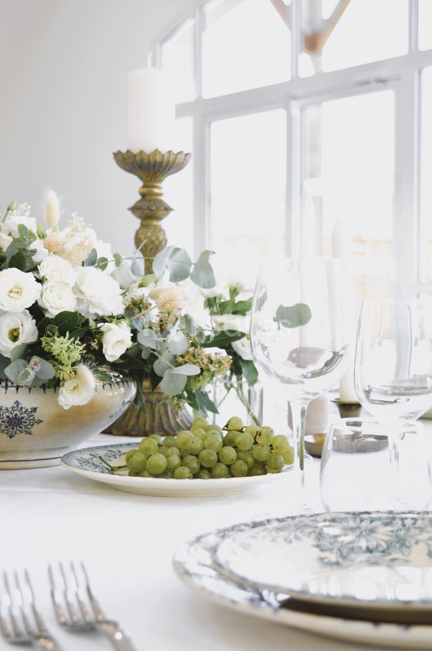 Table élégamment dressée avec assiette de raisins, bouquet de fleurs blanches et chandelier près d'une fenêtre lumineuse.
