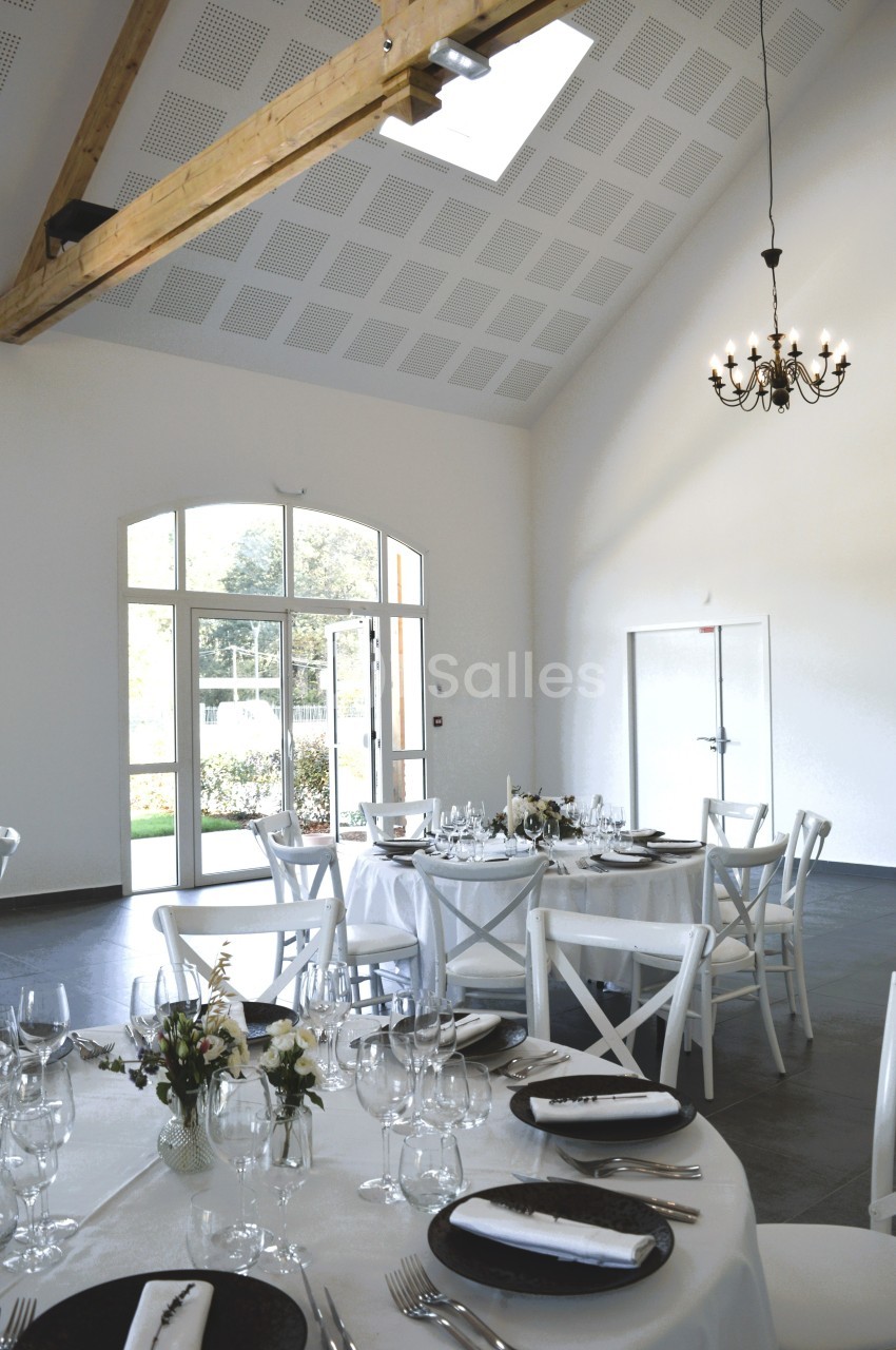 Salle lumineuse avec tables dressées, chaises blanches, poutres apparentes, lustre et grande baie vitrée donnant sur un…