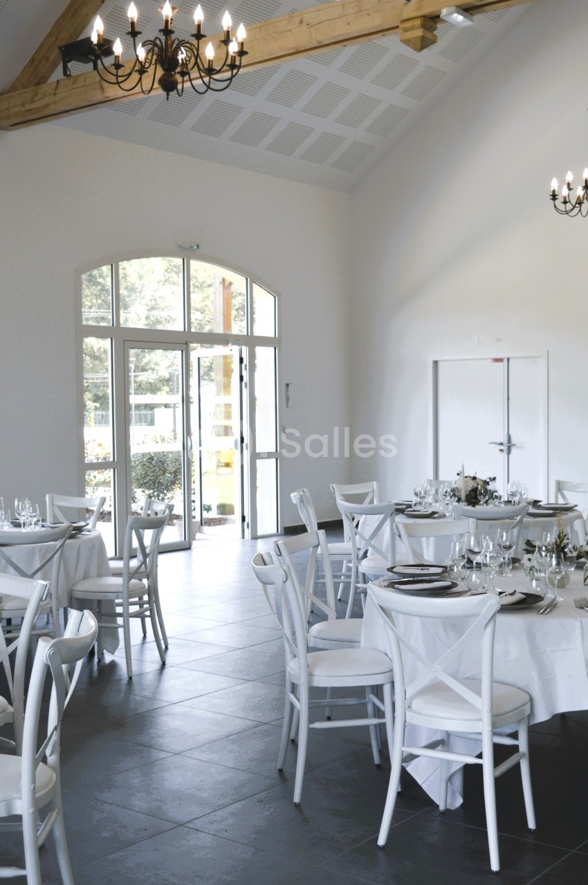 Salle lumineuse avec tables rondes dressées, chaises blanches et grandes fenêtres donnant sur un extérieur verdoyant.