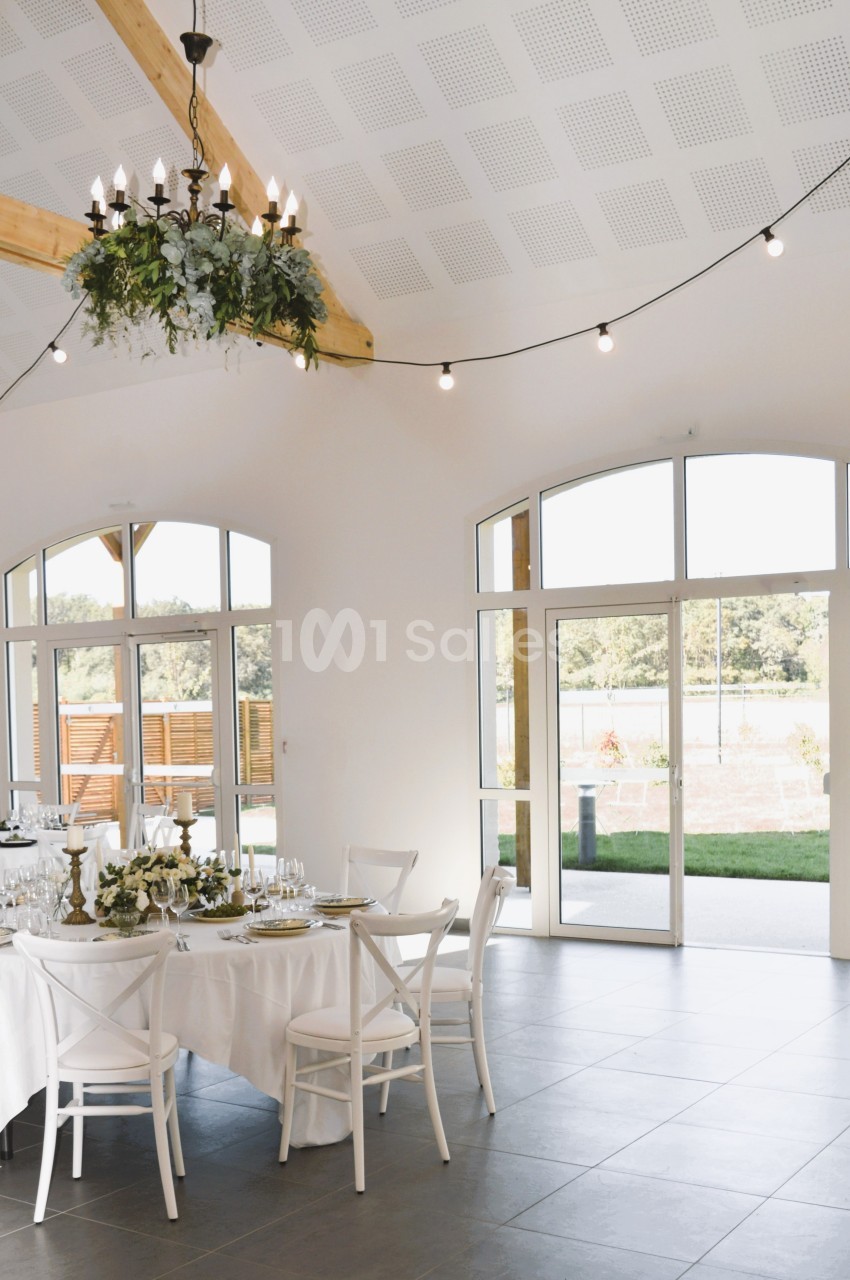 Salle lumineuse avec grandes baies vitrées, tables décorées pour un repas et lustre suspendu au plafond en bois.