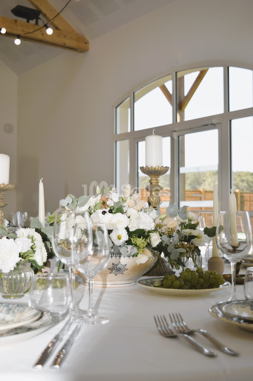Table élégamment dressée avec des fleurs blanches, bougies et assiettes, dans une salle lumineuse avec baie vitrée.