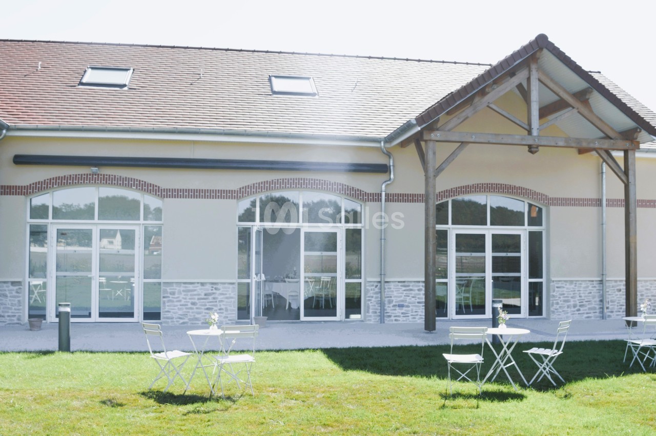 Façade d'un bâtiment moderne avec grandes baies vitrées, terrasse aménagée avec tables et chaises sur pelouse.