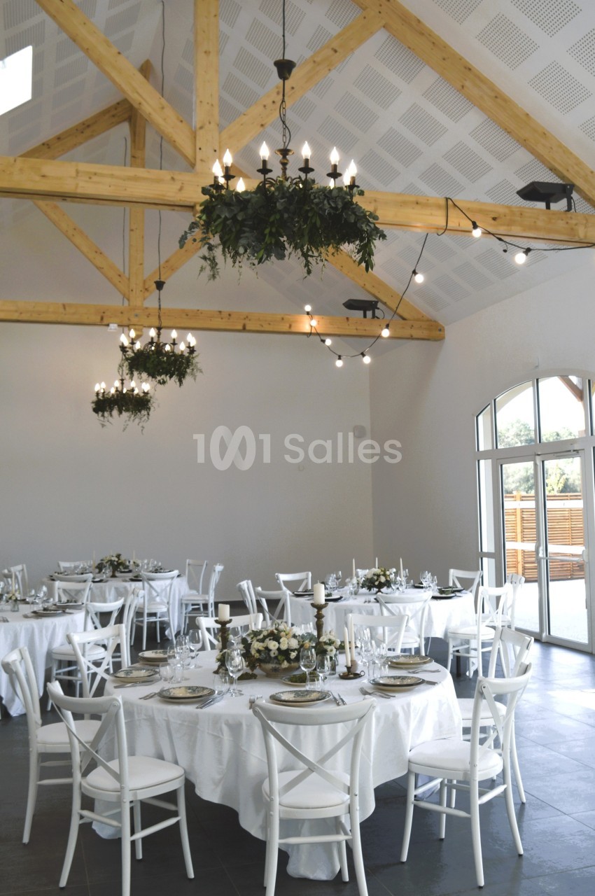 Salle de réception lumineuse avec tables rondes dressées, chaises blanches et poutres apparentes en bois.