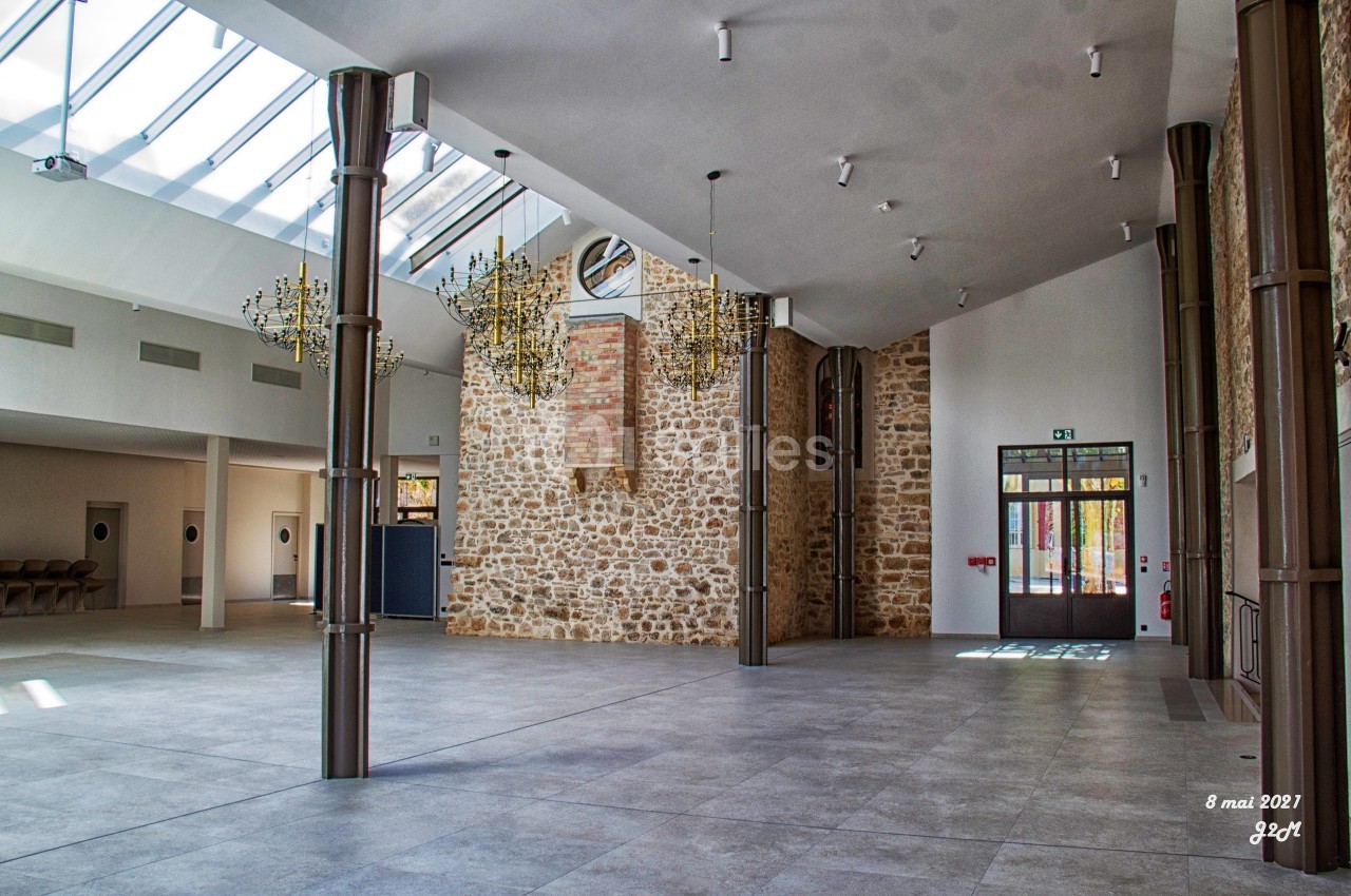 Salle spacieuse avec murs en pierre, grandes baies vitrées, lustres suspendus et sol en carrelage gris.
