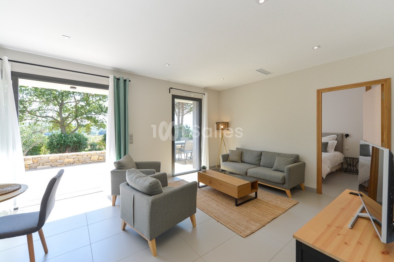 Salon lumineux avec canapé, fauteuils, table basse, ouvert sur une terrasse et une chambre visible en arrière-plan.